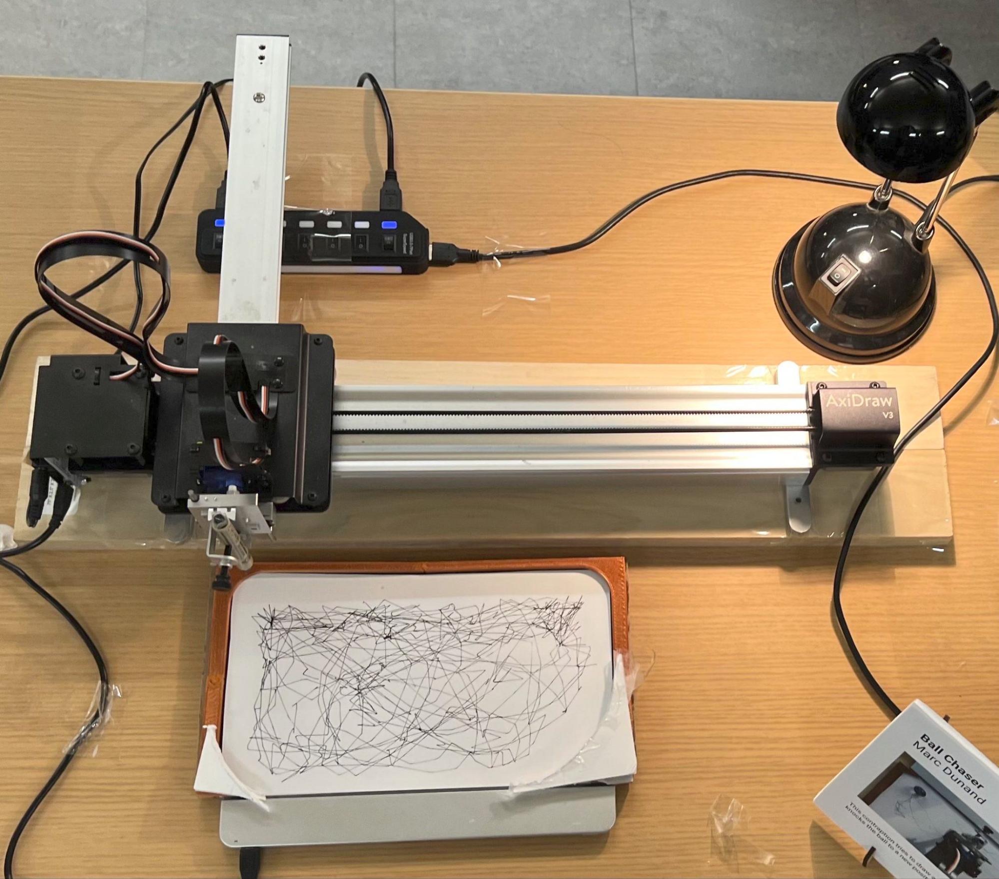 The installed piece on a table: the AxiDraw on a wooden base, the printed arena holding paper over the Sensel pad, a desk lamp, and a printed placard - and no camera anywhere