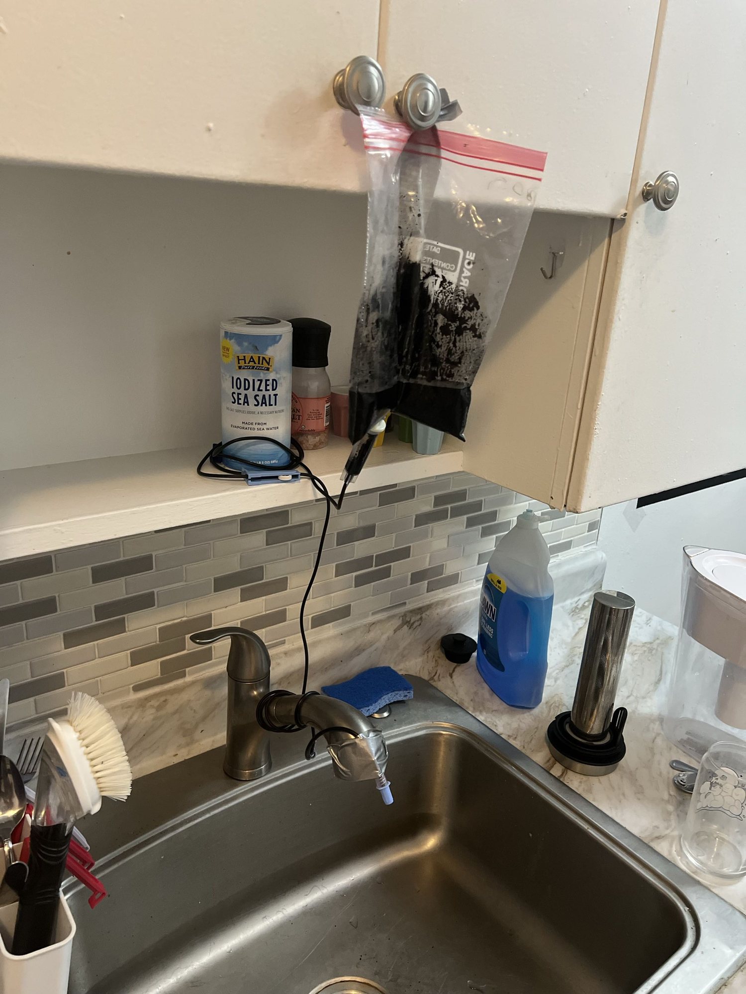 A Ziploc bag of ink hung from a kitchen cabinet above a sink, with a line running down to the faucet