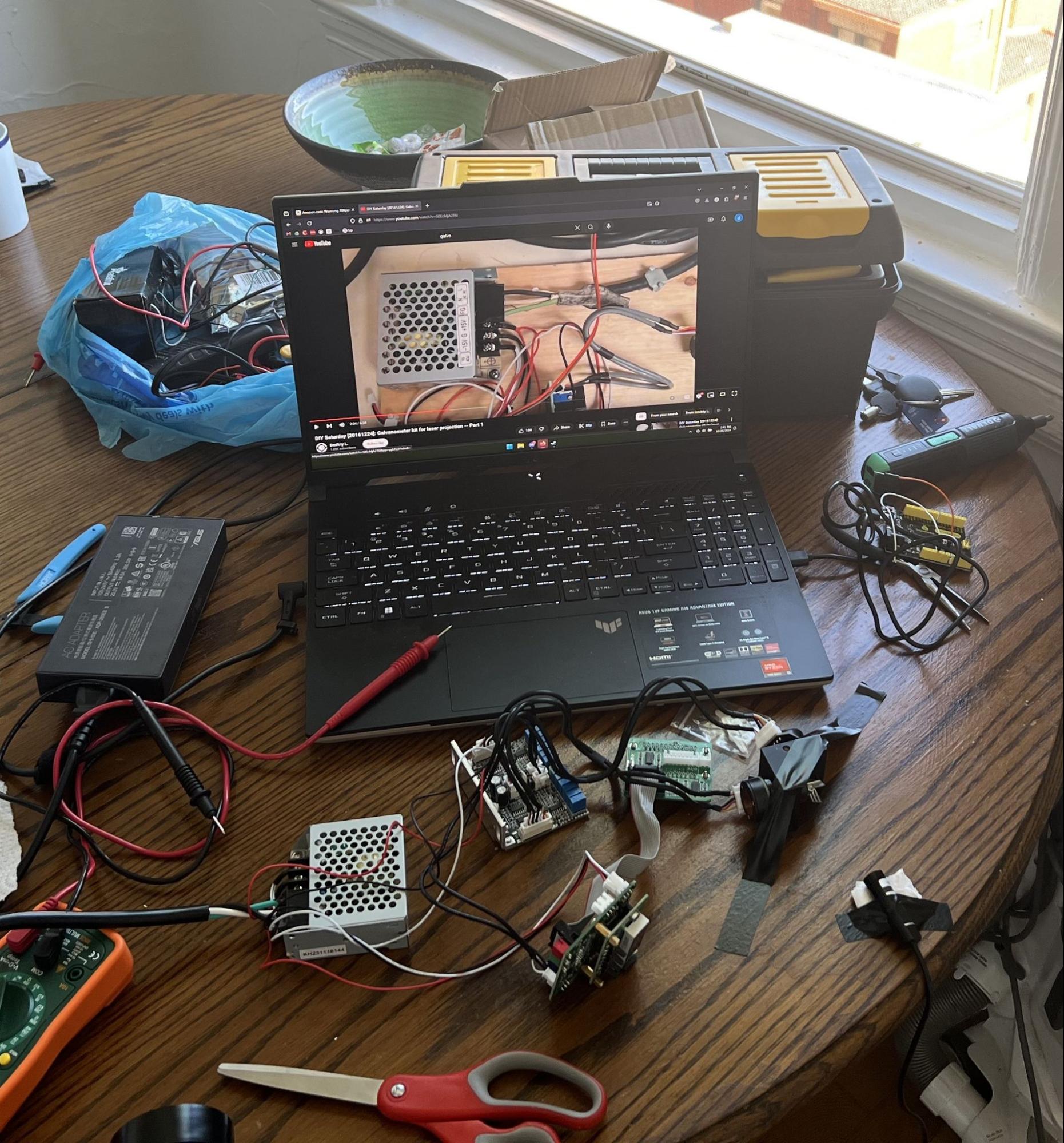 A laptop playing a galvanometer assembly tutorial, surrounded by galvo components, a multimeter, and hand tools